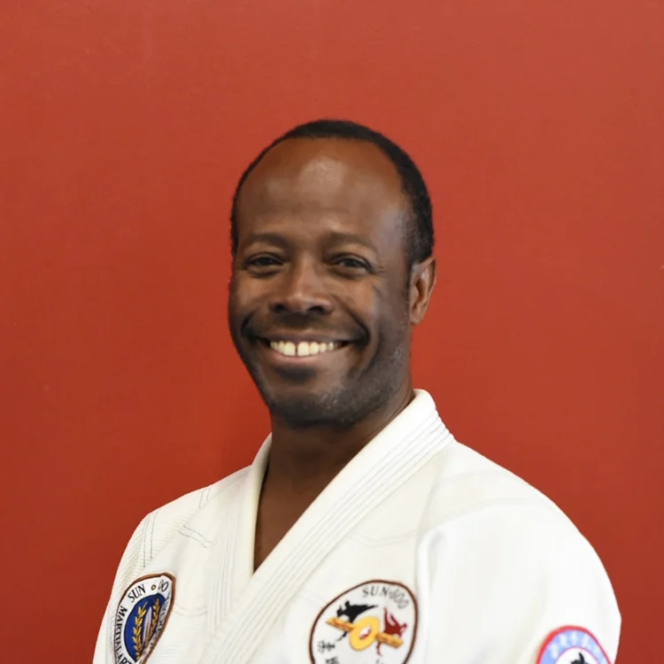 Professor Thabiti Sabahive, wearing a white martial arts uniform with patches, smiles at the camera as he stands in front of a plain red background.