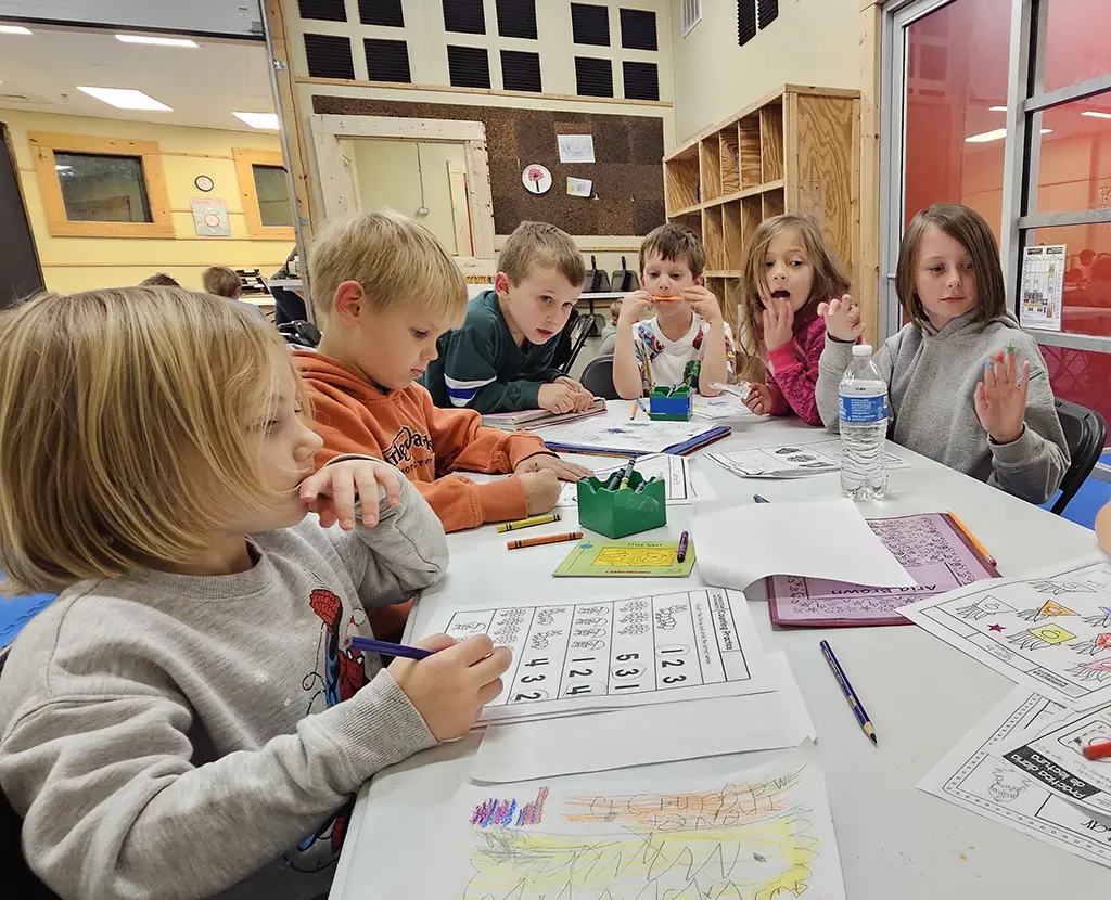 Six young children sit around a classroom table during After School, working on worksheets with colored pencils and crayons. Some are coloring, and others are talking or raising hands. The classroom background has shelves and a corkboard, reflecting the engaging environment at Asheville Sun Soo Martial Arts.